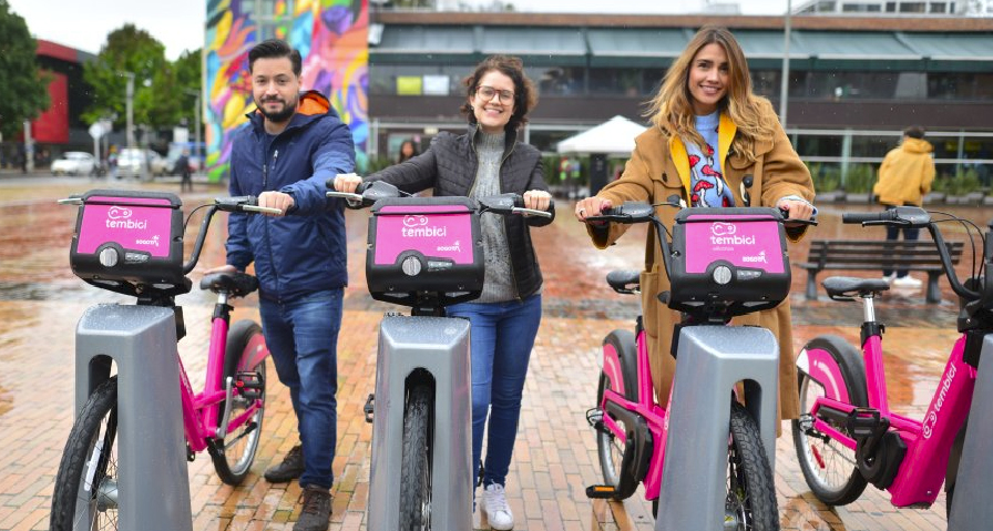 Tres personas, dos chicas y un chico posan con bicicletas del sistema de bicicletas compartidas de Bogotá identificado en rótulo rosa como Tembici