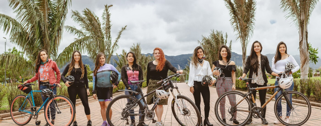 Un grupo de chicas posa mirando a cámara, en una zona peatonal junto a sus bicicletas. Hay vegetación de media altura a ambos lados y al fondo se ve un cielo gris y una cordillera montañosa