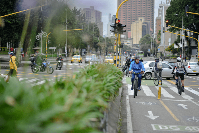 Una avenida de Bogotá. En un cruce con semáforos confluyen ciclistas en uso de ciclovía, peatones, motociclistas y vehículos particulares