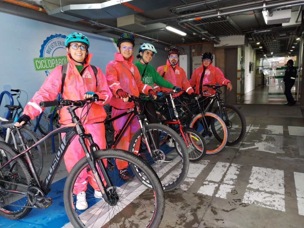 Cinco personas posan con sus bicicletas. Llevan casco y chubasquero de color rojo para protegerse de la lluvia. Están en un parqueadero bajo techo. Se ve al fondo una una oficina.
